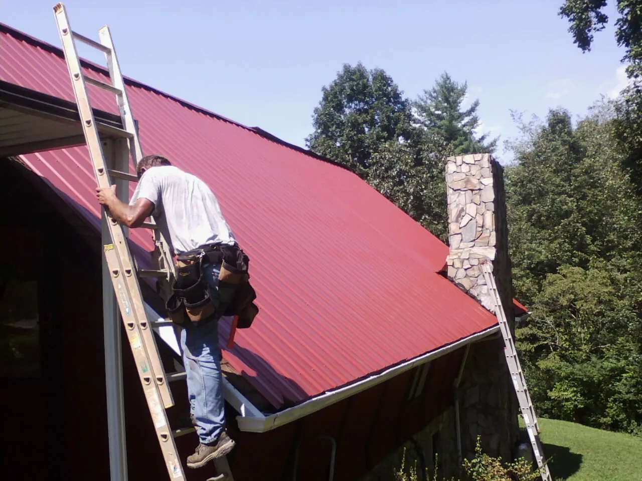 Roofer on a ladder beside a red metal roof and stone chimney — Soffit & Fascia Repair by Roofing Brothers in Liberty
